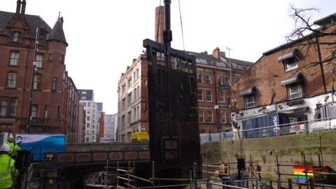 Photograph of the old lock gates being lifted from the Rochdale Canal in Manchester city centre. The image shows a crane, scaffolding and workmen. 