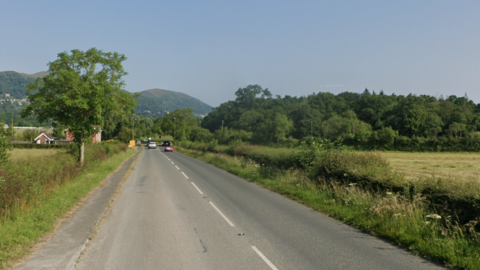 The B4208 road on a sunny day. The road is lined on both sides with hedgerows bordered by trees. A few cars are visible in the distance and beyond them are the Malvern Hills.