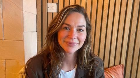 Jenny Morgan smiling at the camera as she sits near a wall of wooden slats. She has dark brown hair with blonde highlights which is framing her face. She has a white t-shirt on with a black leather jacket.