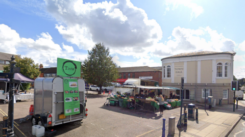 A Google Streetview image of Lutterworth Market Street car park on market day with a food truck and a fruit and veg stall. The food truck has a curved roof and is made from silver metal, with a green sign. The market stall has a green and white canvas roof.