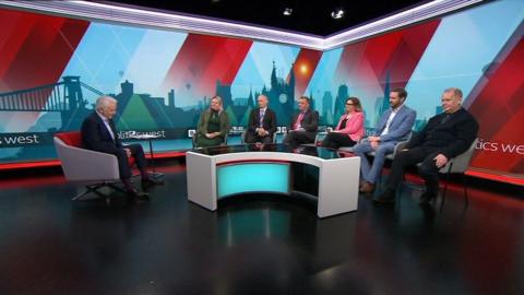 Six of the mayoral candidates sit on chairs in the BBC Politics West Studio. They are all sitting in front of a table. The presenter David Garmston is sitting in a chair to the side of the studio.