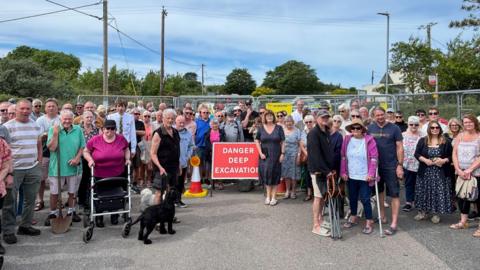 A picture of a group of residents looking forward. They are stood in front of a metal fence with a sign that reads 'Danger Deep Excavation' on it.