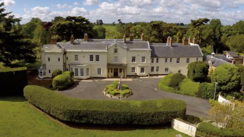 Moulton Grange is a cream coloured building with a grey slate roof and brick chimneys. This view shows the front façade, including an entrance porch with two columns on either side, a bay window to the left of the porch and multiple lattice pane windows. The approach to the property is a wide driveway surrounded by hedges with a circular bush in the centre of the drive and a union flag on a white pole.