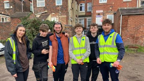A group of five men and one woman stand in a line and smile at the camera. Some in the group wear hi-viz vests. They stand behind a row of red-brick buildings.