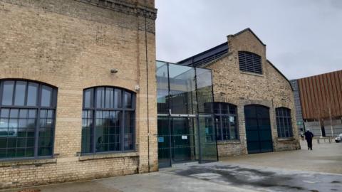 The outside of Sand Martin House in Peterborough. There are two yellow bricked buildings, with a glass atrium in the middle
