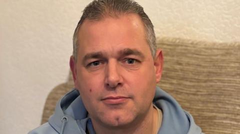A man in a light blue tracksuit top with short hair sitting on a beige sofa looks at the camera unsmilingly 