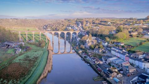 Calstock Viaduct, a bridge comprising tall stone arches, is reflected in a river running from the background to foreground of the image. To the left of the viaduct is the village of Calstock.