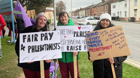 Three female phlebotomists are standing on a grass verge near a roadside holding signs saying "fair pay for phlebotomists" and "behind every diagnosis there is a phlebotomist".