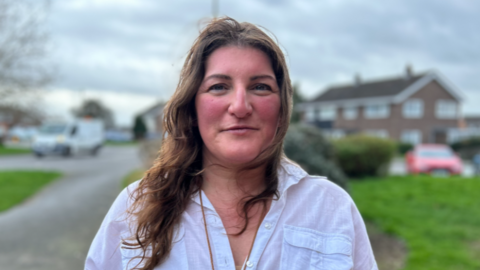 A woman with long brown hair and a white blouse in front of a blurred street with grass verges