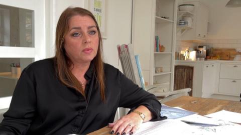 Resident Lisa Miller has long dark brown hair and is wearing a black shirt. Her nails are painted black. She is pictured sitting at a kitchen table covered with maps, plans, and letters. 