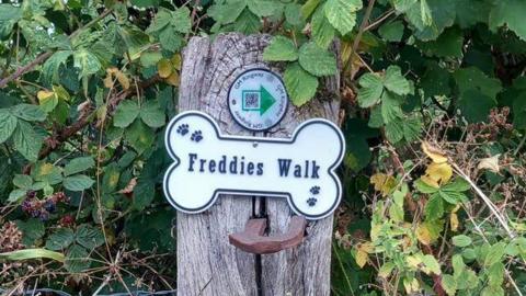 On a wooden signpost, there is a white sign in the shape of a dog bone with the words "Freddies Walk" surrounded by black pawprints. There is also a smaller circle sign above it saying GM (Greater Manchester) Ringway. Green leaves and shrubs form the backdrop.