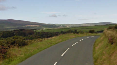 A stretch of road between a grassy bank and green fields, with hills in the background.
