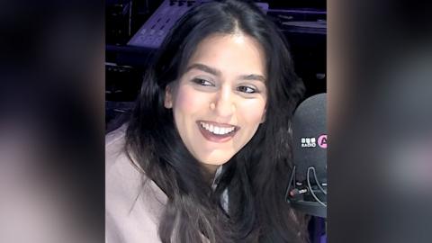 A woman with long, dark hair smiles as she talks into a microphone