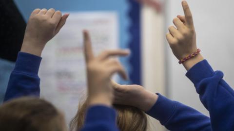 A picture of children sat in a classroom with their hands up. They are wearing blue jumpers.