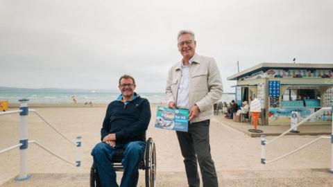 Two men are pictured together atop a short staircase that leads to a beach. The man on the left is wearing glasses and sitting in a wheelchair. The man on the right, wearing a cream jacket, is holding a sign that has 'A Coast for All' printed on it. Behind them, and to the right, there is a small building selling cream. 