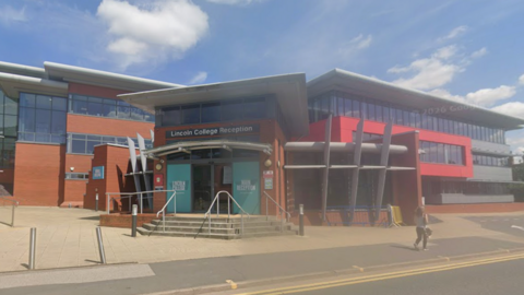 The Lincoln College reception building. It is a red brick multi-storey building with lots of windows. It has double doors leading inside the building. The sky is blue with some white clouds. 