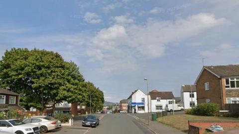 A residential street, houses with large trees are seen to the left and more houses to the right of the street