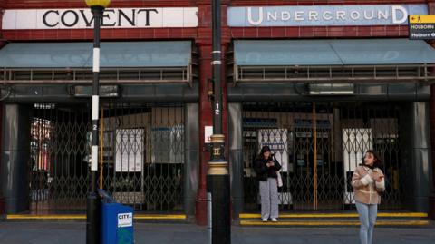 Women on phones stand outside Covent Garden Tube station with shutters closed