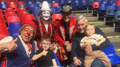 A man and two young children sit with three performers from the circus. One performer, dressed as a clown has a red nose, a red hat and is wearing a blue waistcoat. Another performer is wearing a red costume with a white face paint while a third performer is dressed as the ringmaster in a red suit, black shirt and a black hat with gold detailing.