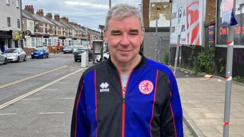 Robert Nichols standing on Linthorpe Road. He has short grey hair, brushed to his right, and is wearing a blue-and-black Middlesbrough FC tracksuit top. A row of terraced houses can be seen in the background on the left, while on the right are two advertising billboards.