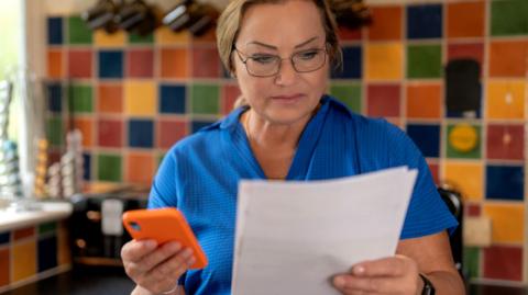 Woman in a kitchen holds a smartphone in one hand and a bill in the other, with colourful tiles on the wall behind her.