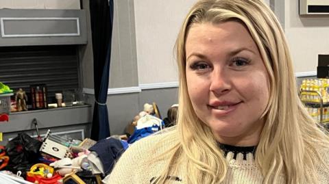 Katie Symmons standing in a large hall. Behind her are piles of donated goods, including tinned food and clothing.