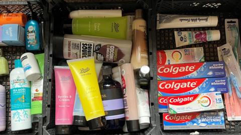 Three plastic boxes of hygiene products have been sorted so in one box there are various brands of shampoo and conditioner. The middle box has various types and brands of shower gel while in the last box there are dental products including tubes of toothpaste, children's toothpaste and toothbrushes.