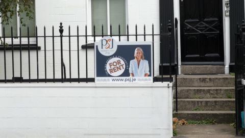A For Rent sign on a residential terraced house, with a picture of a blonde woman in a white suit. The house is white with black railings and steps leading up to a black door.