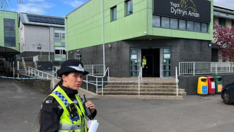 Female police after stood in front of the front entrance to Ysgol Dyffryn Aman with police tape at the school entrance and another uniformed officer in the background