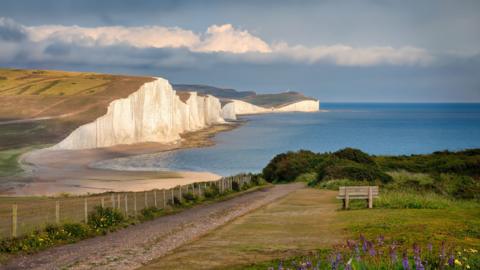 Picturesque white cliffs, known as the Seven Sisters, face a pale blue ocean. The sun is setting. 