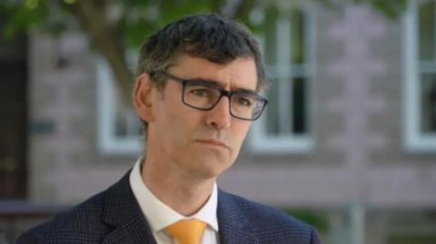 A picture of a man wearing black glasses looking behind where the photo has been taken. He is wearing a navy suit jacket, a white suit shirt and a yellow tie.
