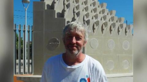 Rob Macdonald is standing in front of the Solidarity Park living monument in Spain. He is wearing a white t-shirt with a Solidarity Park logo on it and has white hair and a beard. The sculpture is made of white stones and resembles a hip. 