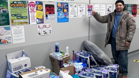 Glenroy Bell stands before a board pointing at a poster hanging on it. He is wearing a grey coat and blue jumper and jeans. Beside him are a number of packets of toilet roll.  