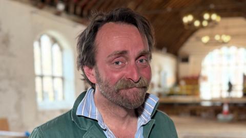 Tim FitzHigham wearing a green jumper over a blue and white shirt. He has brown hair and a thick moustache and is smiling while standing inside the Guildhall, which has long windows and chandeliers hanging from the ceiling.