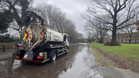 A white road tanker with a tube attached to the back of it. The lorry is parked on a flooded road with a patch of grass and trees to the side.