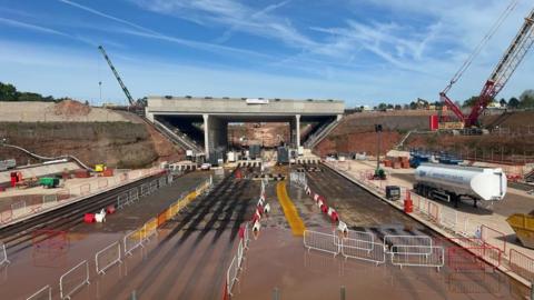 Construction work taking place on an A road to install a bridge. Cranes and barriers can be seen against the backdrop of a blue sky.