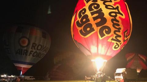 A large creme egg branded hot air balloon being inflated at night, so it looks like it is glowing. The flame from the burner is visible and there are other hot air balloons tethered on the ground in the background