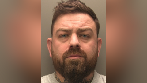A police custody shot of Max Skidmore looking at the camera. He has short brown hair and short brown facial hair. He has tattoos on his neck and is wearing a grey sweatshirt.
