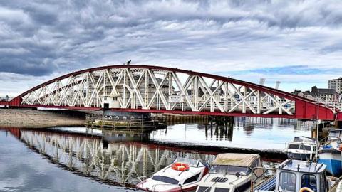 Ramsey Swing Bridge, a curved red and white structure with white x-shaped metal bars.The water is flat calm and there are a number of boats in the foreground. 