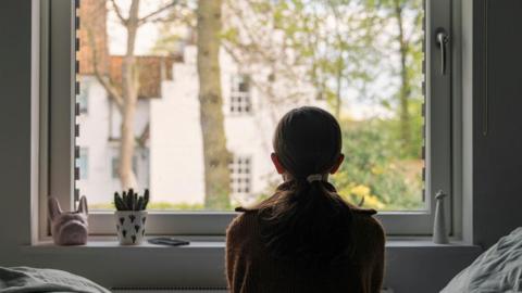 A young girl looks out of her bedroom window. The view through the window is blurry but shows a residential area.