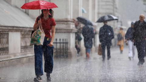 A woman with a red top, red umbrella walks with shopping down a street in the rain. There are other figures in the background.