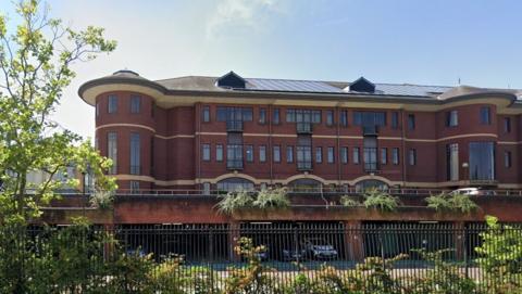A large red-brick office building with rounded corner towers and a grey roof stands behind a black metal fence and landscaped shrubs. The three-storey building features rows of windows and balconies, with solar panels visible on the roof. Trees and greenery frame the scene under a clear blue sky.