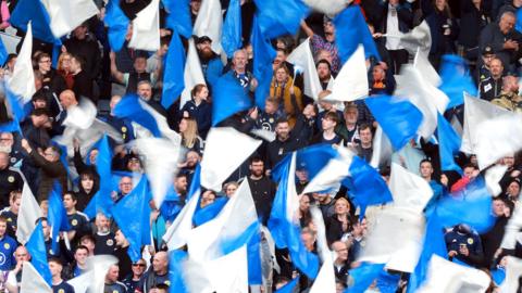 Football fans wave blue and white Scotland flags while watching the Scotland v Finland international friendly being played in Glasgow in 2024.