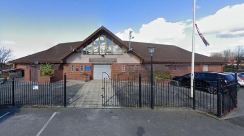 A building with a triangular facade with a sign saying Eldonian Village Hall. A flagpost bearing the British flag is outside in a parking area