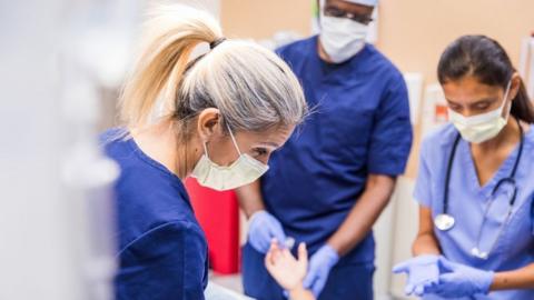 Three medical professionals stood over a patient, they are photographed from waist level up wearing navy scrubs and face masks. In the foreground is a woman with blonde hair in a ponytail. A dark haired woman stands beside her, and a male is in the background holding the patient's hand
