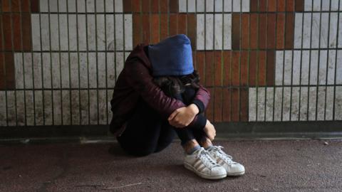 A teenage girl in black jeans, dirty white trainers, a purple jacked and blue hoodie sits of the floor of an underpass, slumped with her head in her lap and arms wrapped around her knees.