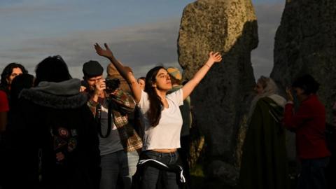 A woman closing her eyes with her hands up, standing inside the stones at Stonehenge