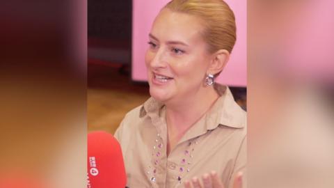 A woman with light ginger hair pulled back from her face talks to someone off-camera.
