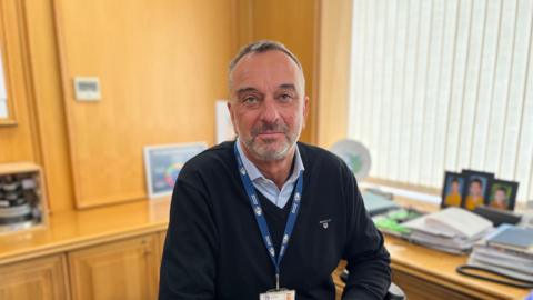 A man with grey and black hair and stubbly grey and black beard sits at a wood-panelled desk with family photos behind him. He's wearing a blue jumper, a blue collared shirt and a lanyard which says Guernsey Electricity on it. 