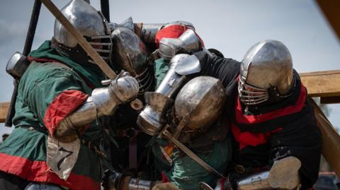 Four people wrestling in medieval-style armour and with swords in hand. Two are in green and red and another two are in red and black clothing with large metal helmets and slatted visors